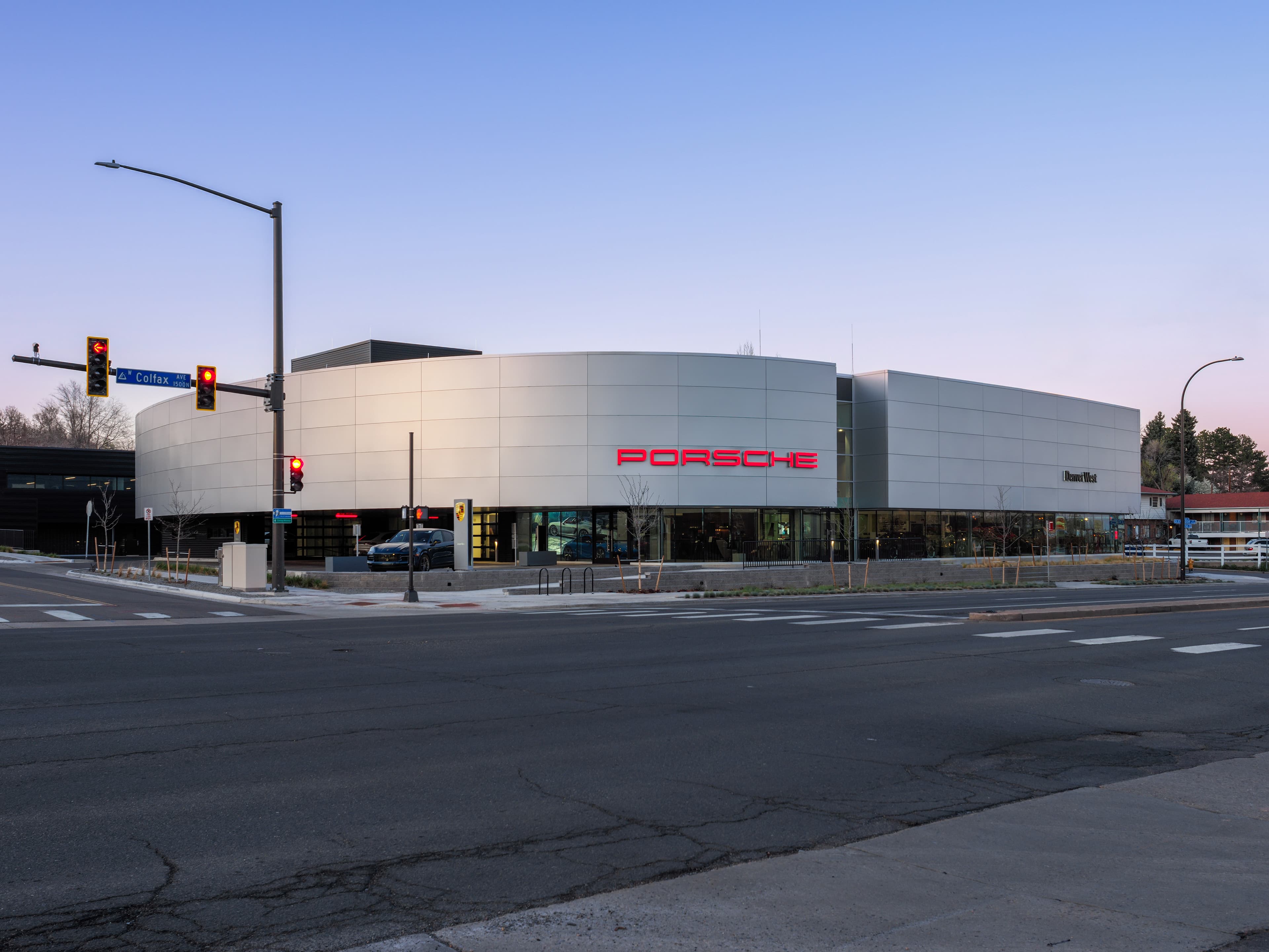 Modern Porsche dealership with sleek design, large windows, and prominent logo, located at a street intersection with traffic lights.